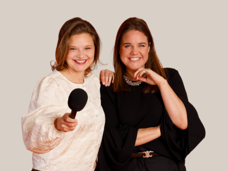 Two women smiling, one with red hair tied up, wearing a floral shirt, the other with brown hair, wearing a black blazer and a cream-colored scarf, against a dark blue background.