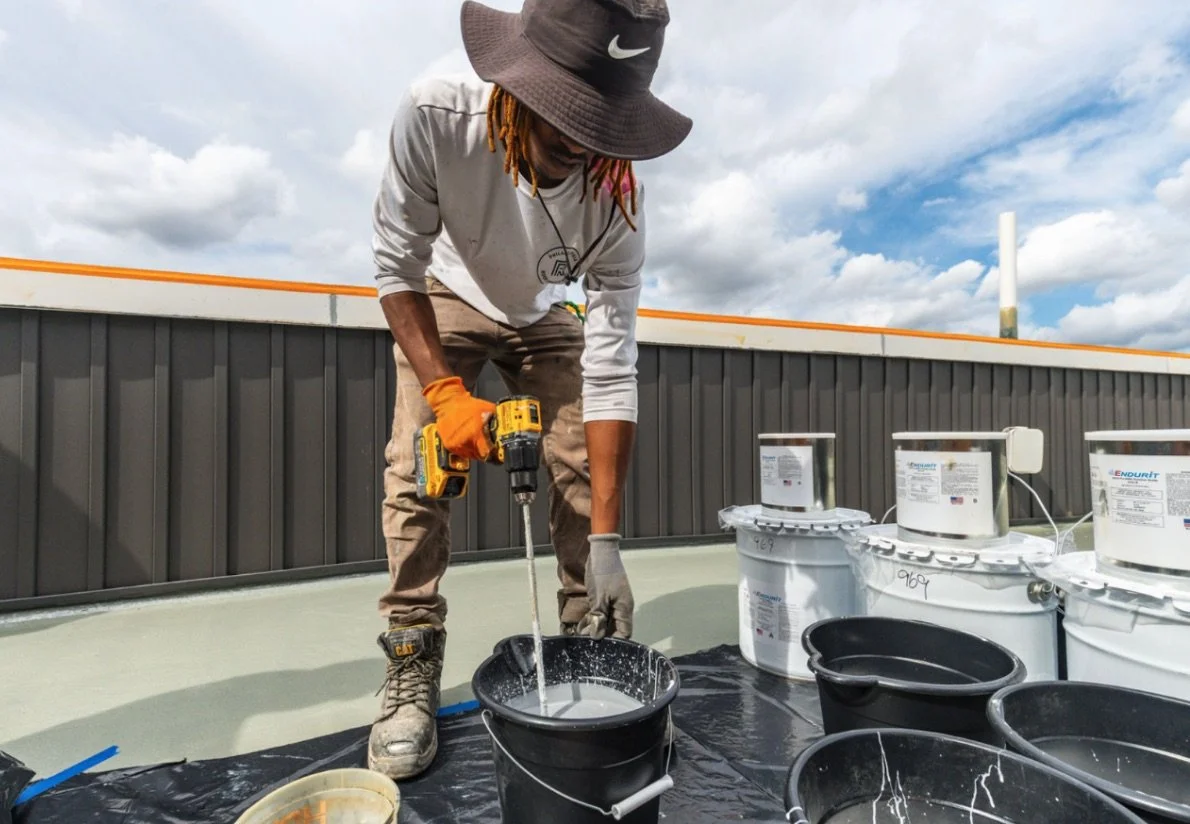 A person working on a roof, using a cordless drill in a bucket with other buckets and cans around, under a partly cloudy sky.