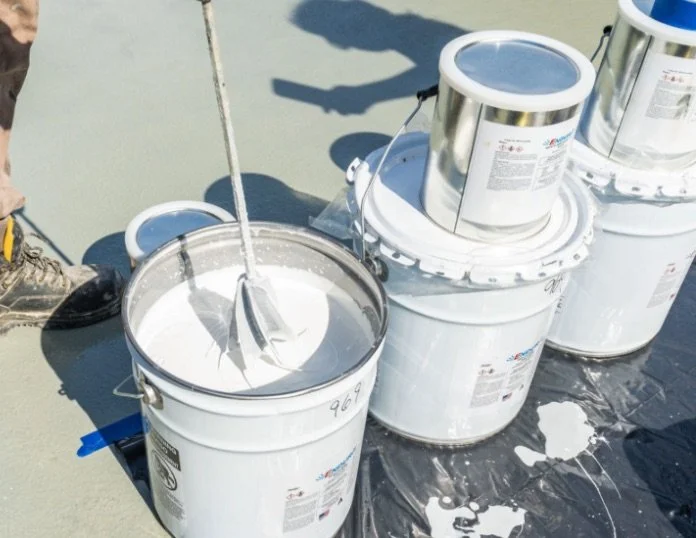 Buckets of white roofing sealant or coating with brushes and a worker's foot nearby.