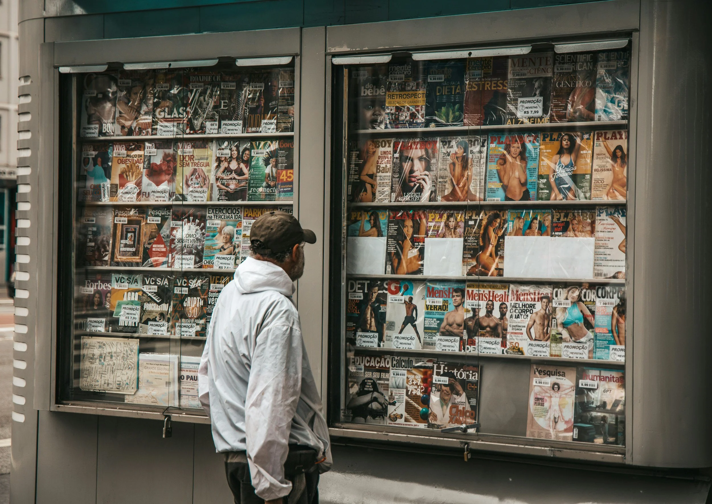 A man standing in front of a newspaper and magazine stand on a sidewalk, browsing magazines displayed behind glass.