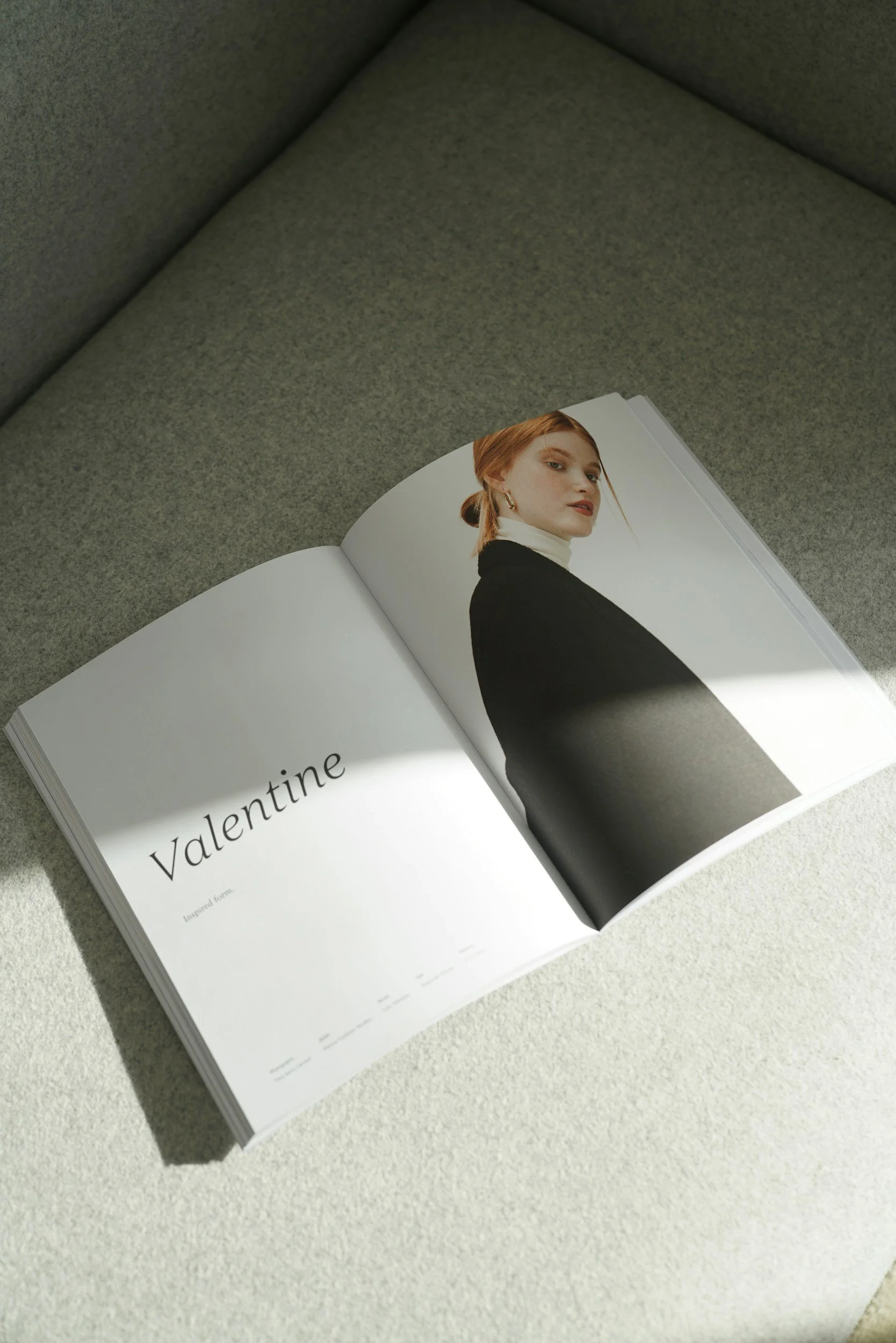 Open fashion magazine on a light-colored textured surface, with a photo of a woman with red hair in a black and white outfit on the right page and the word "Valentine" on the left page.