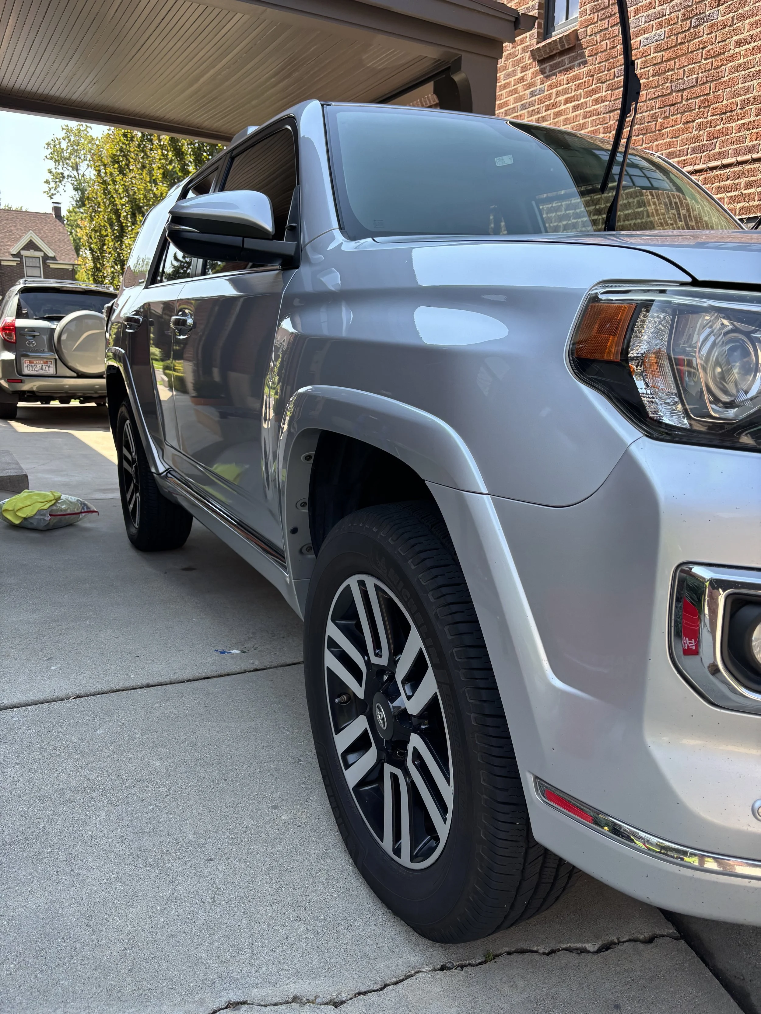 Silver SUV parked in driveway with one of its wheels turned slightly to the left, under a covered parking area outside a brick house.