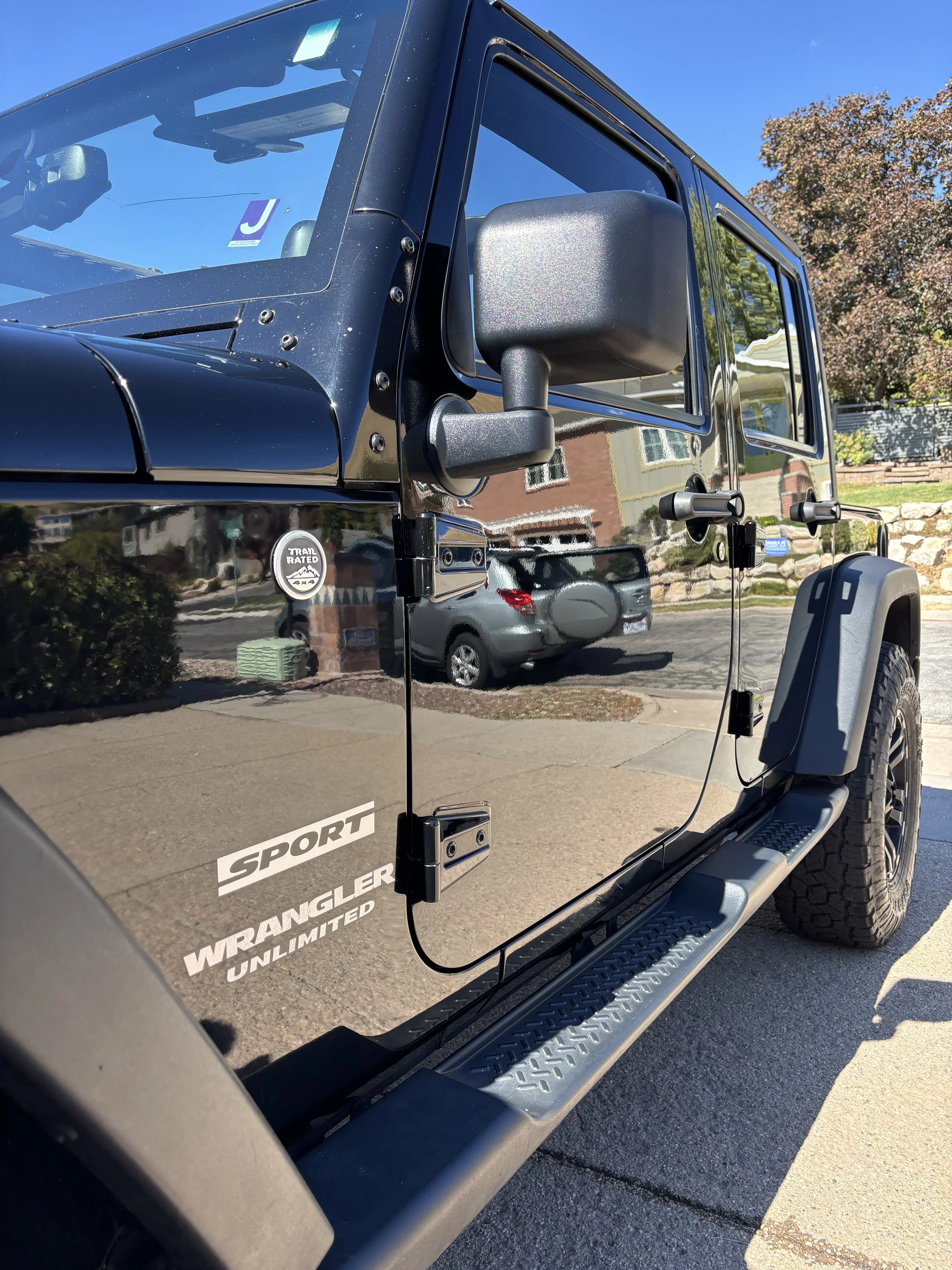 Black Jeep Wrangler Unlimited Sport with side mirrors, tire fender, and window reflections, parked outdoors on a sunny day.