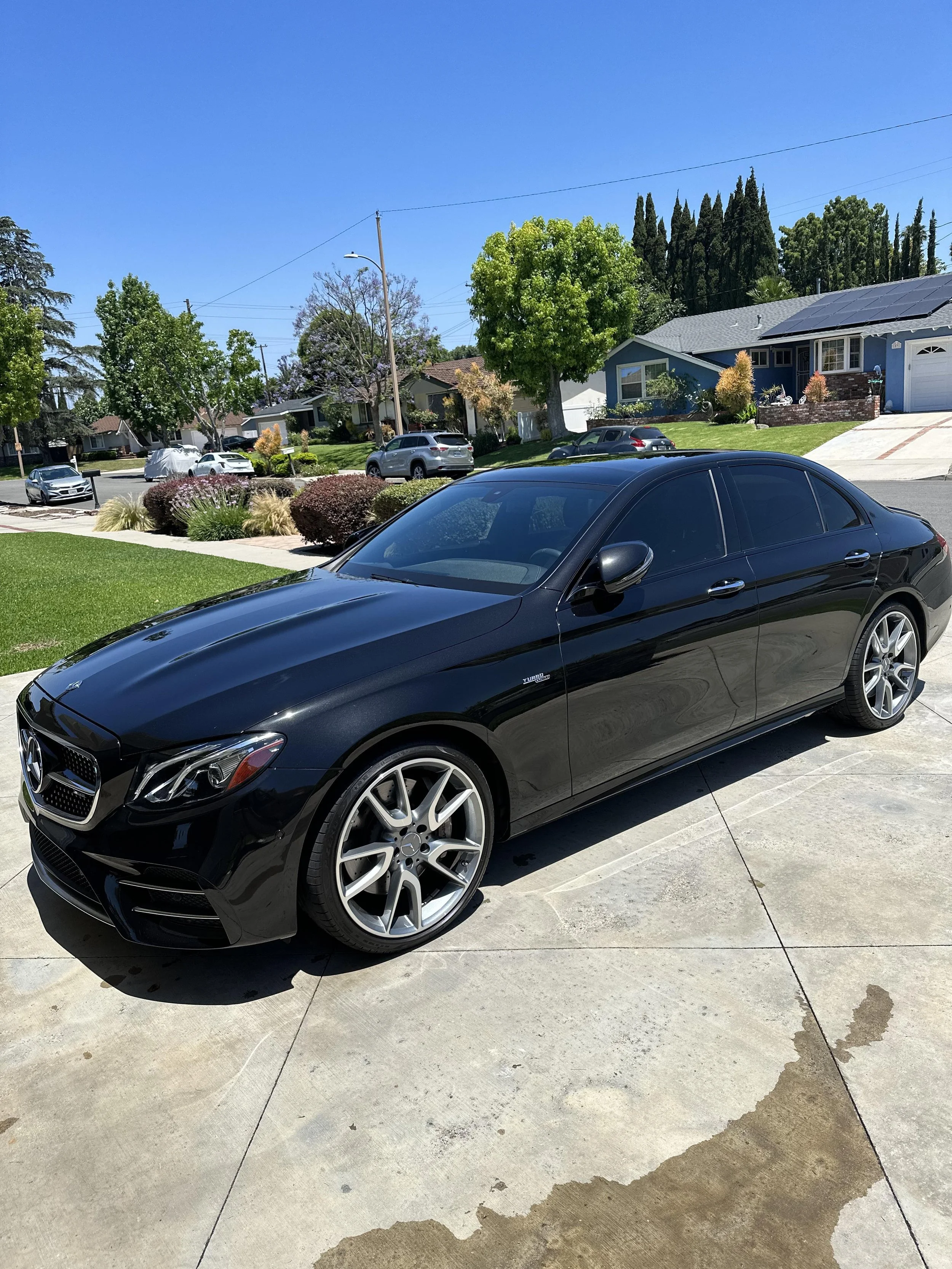 A black Mercedes-Benz sedan parked on a concrete driveway in a suburban neighborhood under a clear blue sky.