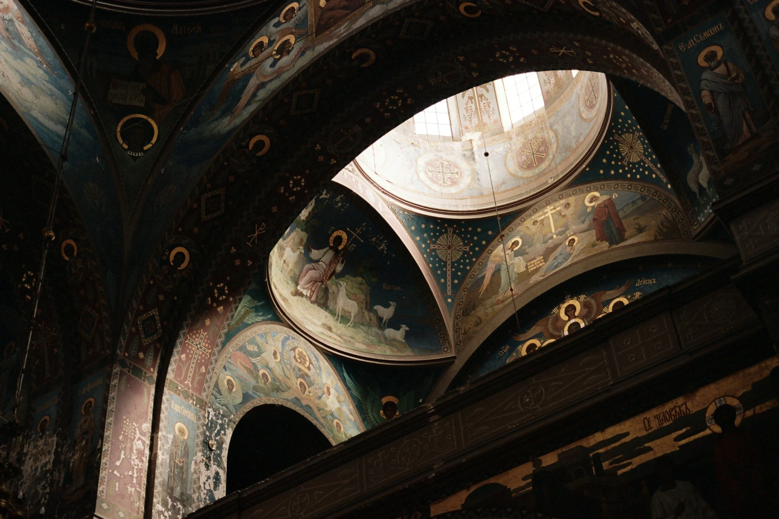 Interior of a church with ornate blue and gold religious murals and icons, including halos around saints' heads, a domed ceiling with a window allowing light inside, and decorative patterns along the arches.