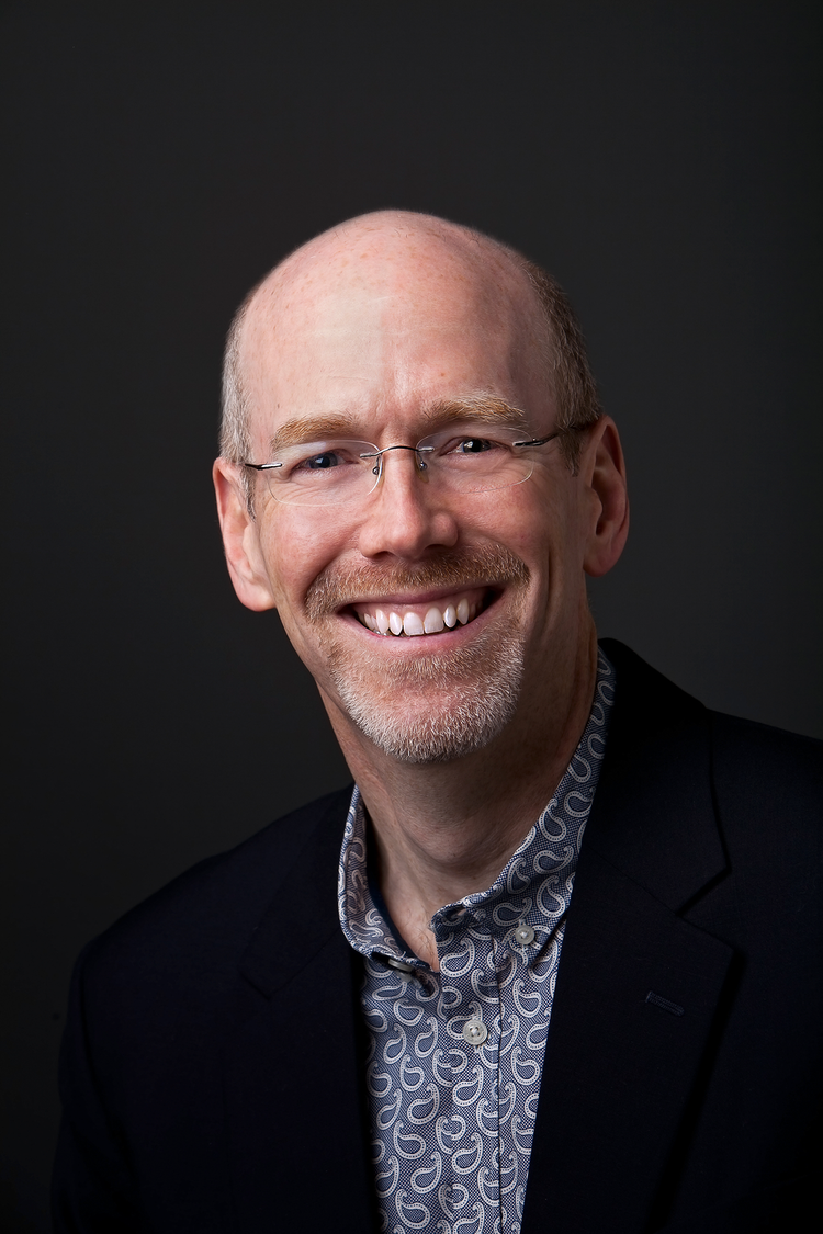Headshot of a smiling man with glasses, a patterned shirt, and a dark blazer against a dark background.