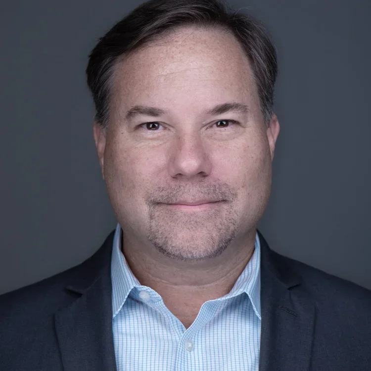 Headshot of a middle-aged man in business attire with short brown hair and slight beard, smiling against a dark background.