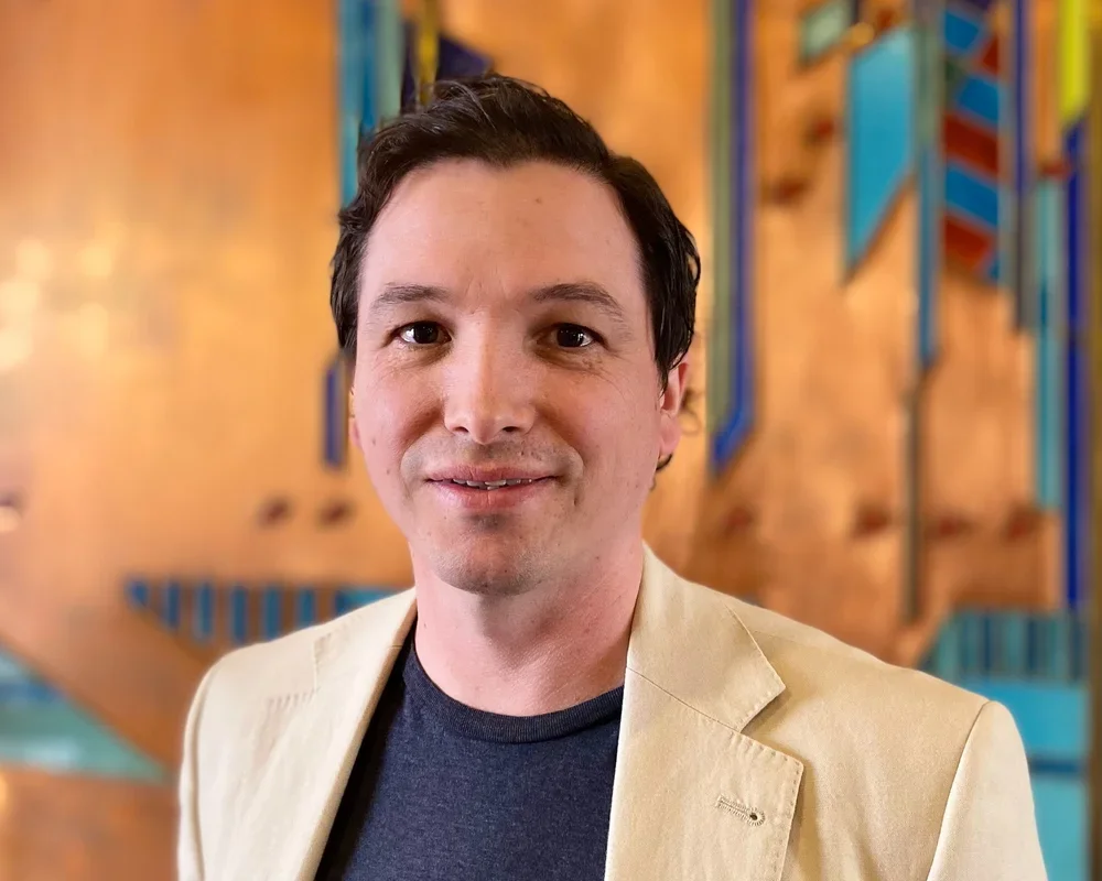 A smiling man with dark hair wears a beige blazer and a navy shirt, standing indoors against a colorful, abstract wall art background.