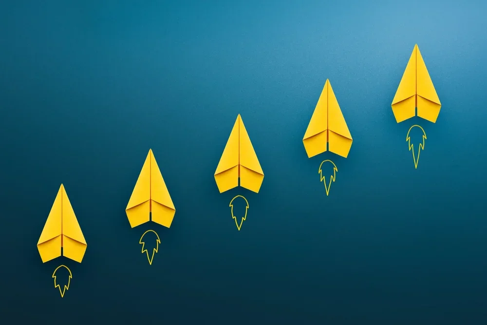 Five yellow paper airplanes arranged in a diagonal line on a blue background, with rocket ship doodles beneath each airplane.