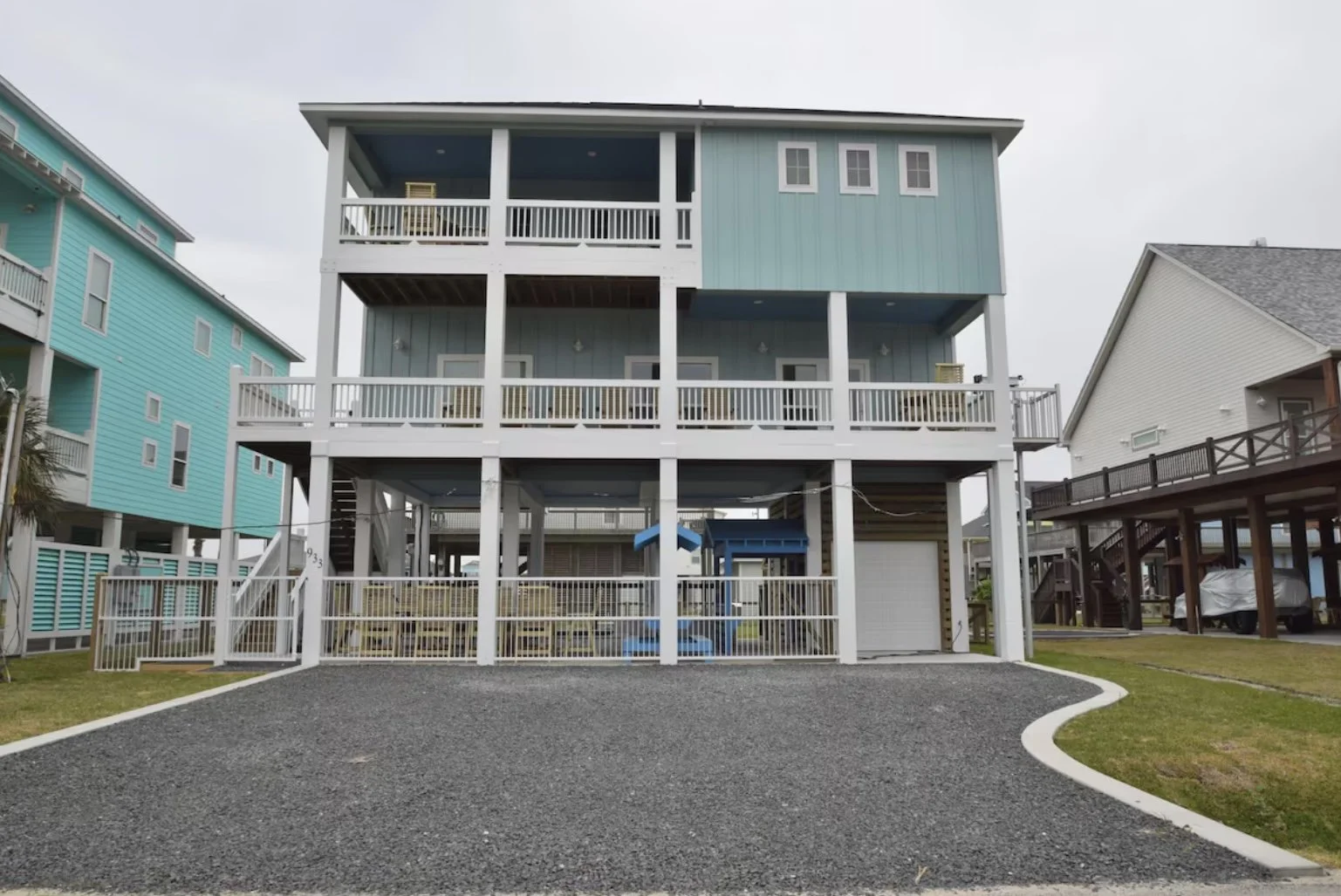 Three-story blue beach house with white railings, open balconies, and a fenced yard, adjacent to other colorful beach homes.
