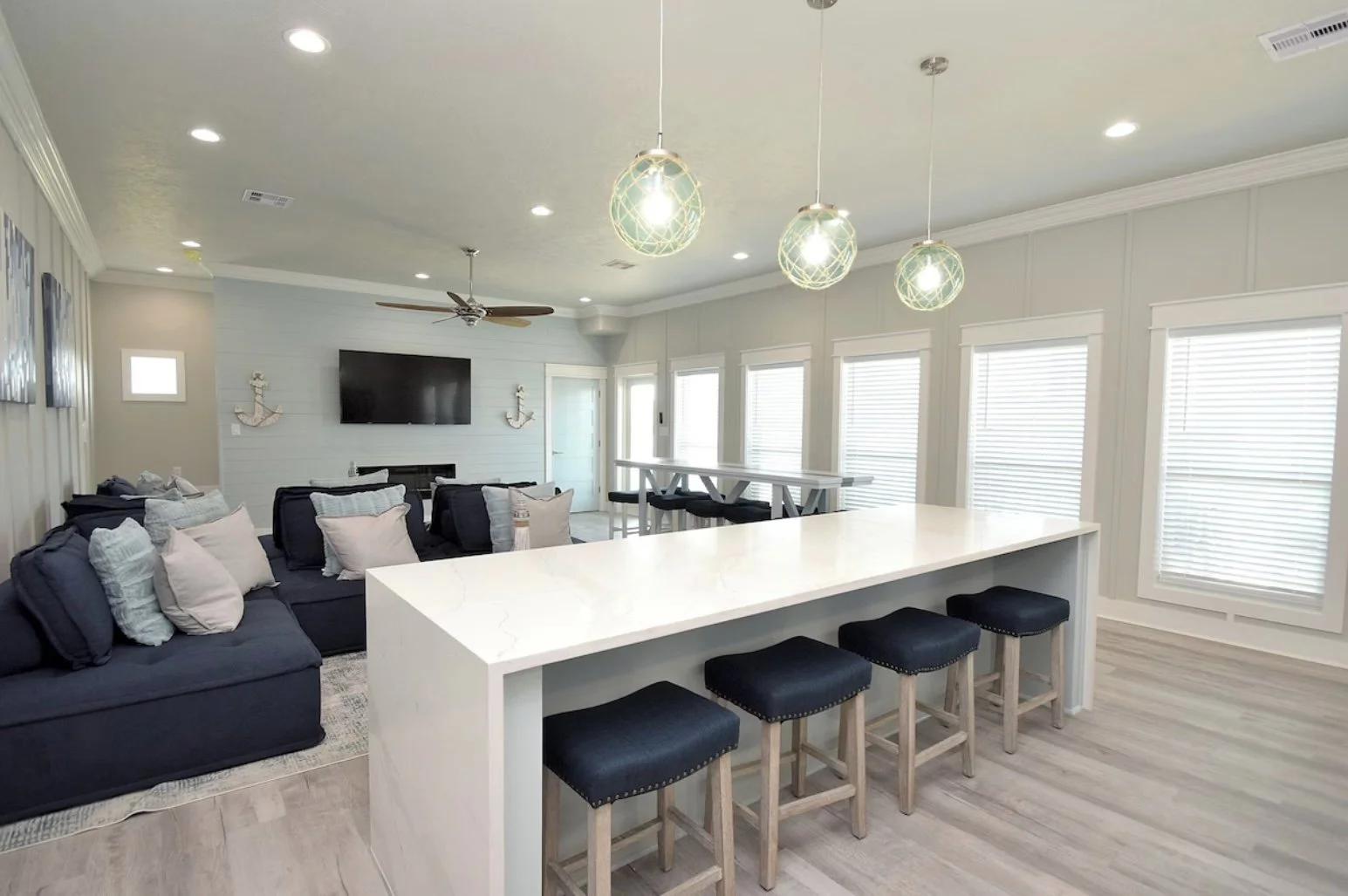 Bright, modern living room with light-colored walls, ceiling ceiling fan, wall-mounted TV, navy sofa with multiple pillows, white kitchen island with four black stools, several windows with white blinds, and nautical-themed decor on the walls.