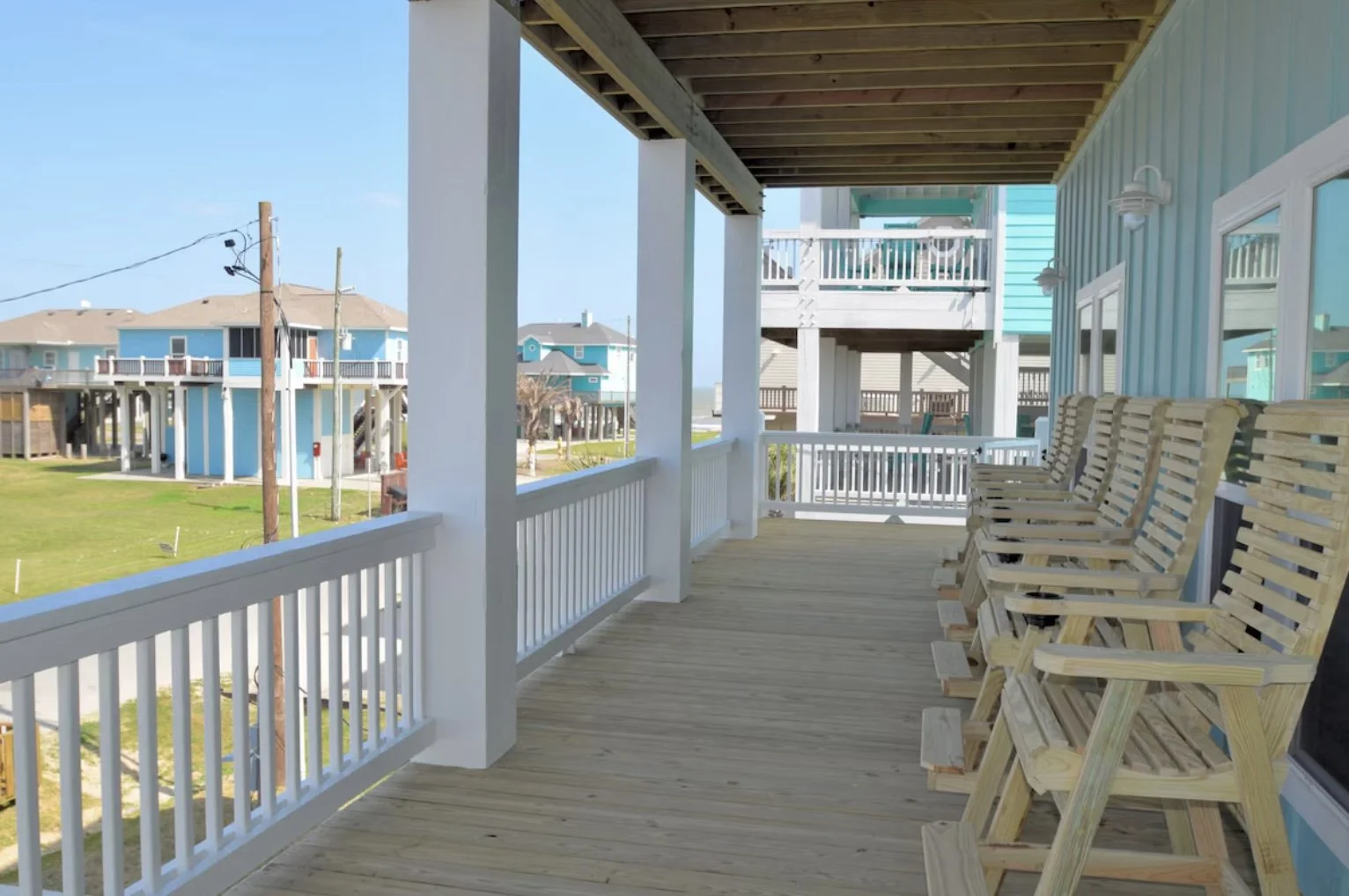 View of a porch with wooden chairs and white railings overlooking neighboring beach houses in a coastal area.