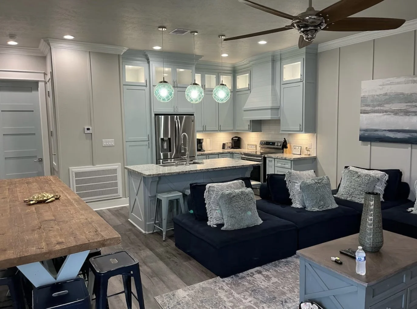 Open-concept kitchen and living room with light gray cabinetry, granite countertops, stainless steel refrigerator, oven, microwave, coffee maker, and a kitchen island with a sink. The living area has a dark blue sectional sofa with gray and silver pi