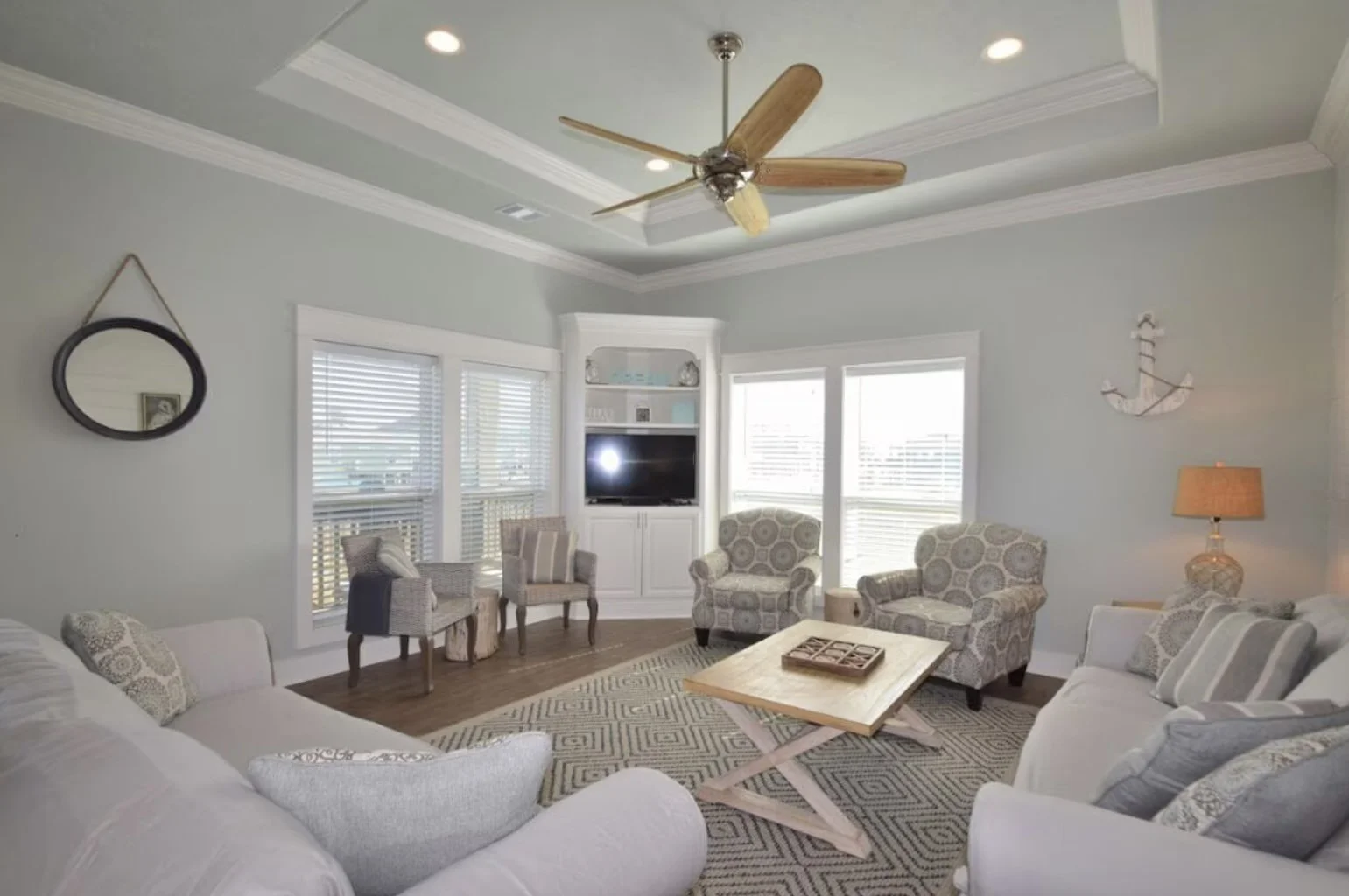 Living room with light gray walls, white crown molding, and a ceiling fan. Furniture includes a beige sofa, patterned armchairs, a wooden coffee table, and side chairs. Decor features a round mirror, anchor wall art, and a lamp. Windows have white bl