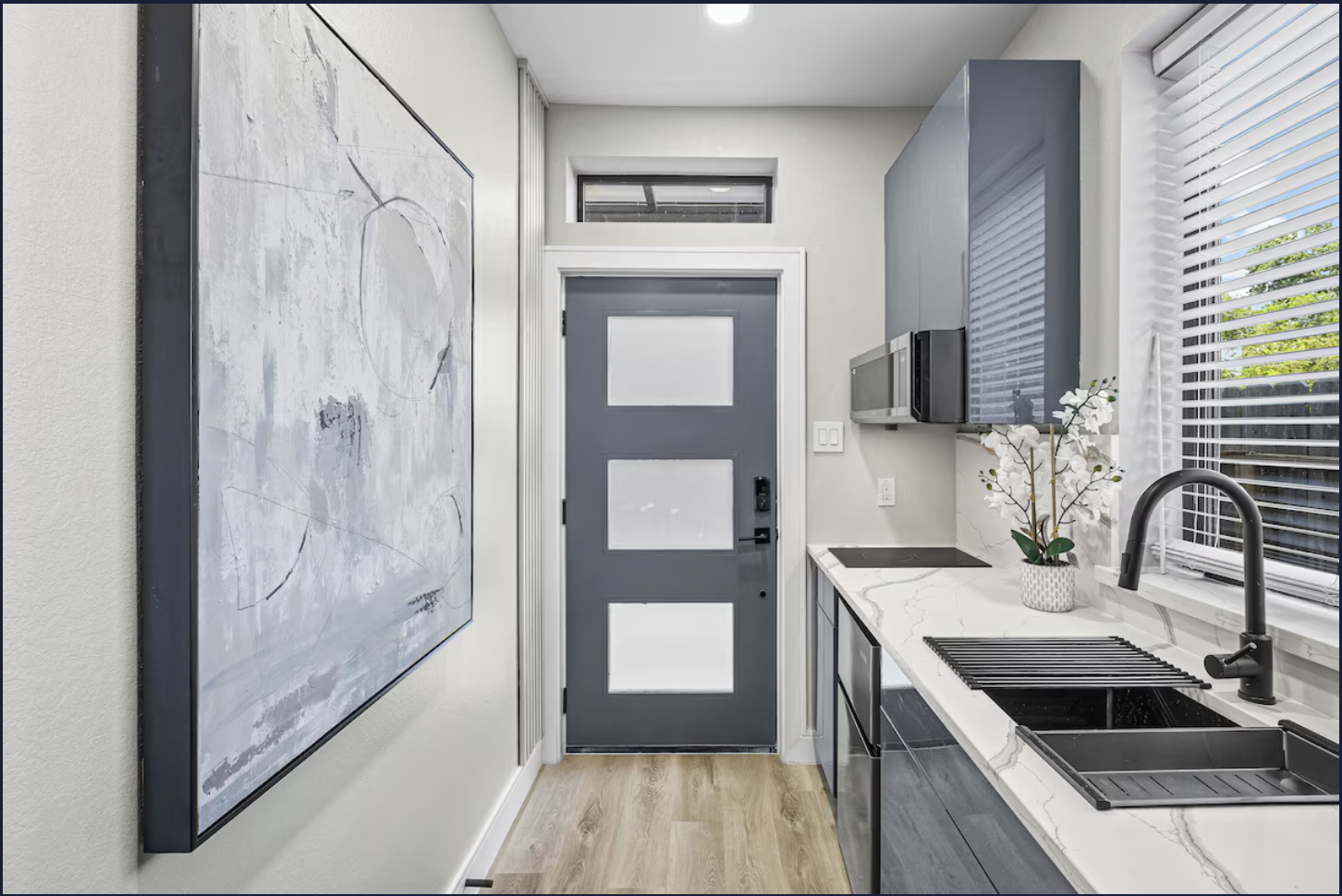 Interior view of a modern kitchen with a gray door, white and gray cabinets, a black sink, a marble countertop, and a window with blinds, a potted plant, and abstract artwork on the wall