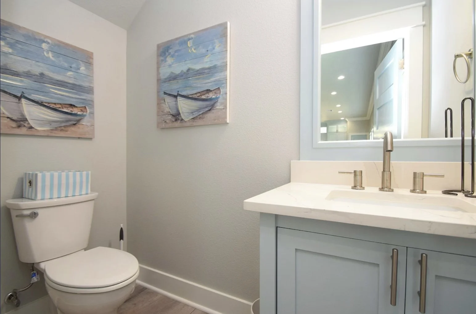 A bathroom with two pictures of boats on the wall, a toilet with a striped tissue box on top, and a light blue vanity with a white countertop and a large mirror.