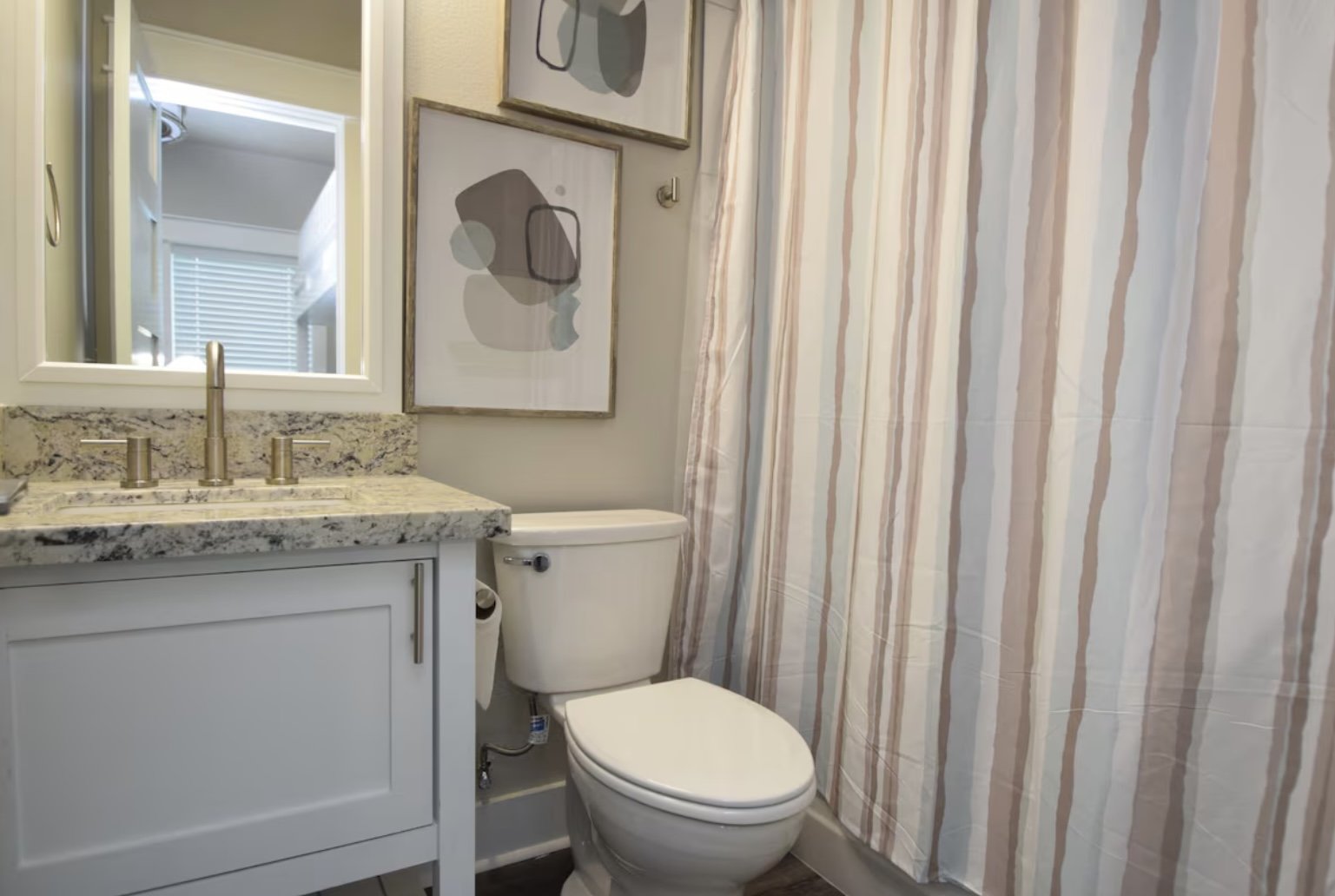Bathroom with a white vanity, granite countertop, and a mirror. There is a toilet and a striped shower curtain. Two abstract artwork pieces hang above the toilet.