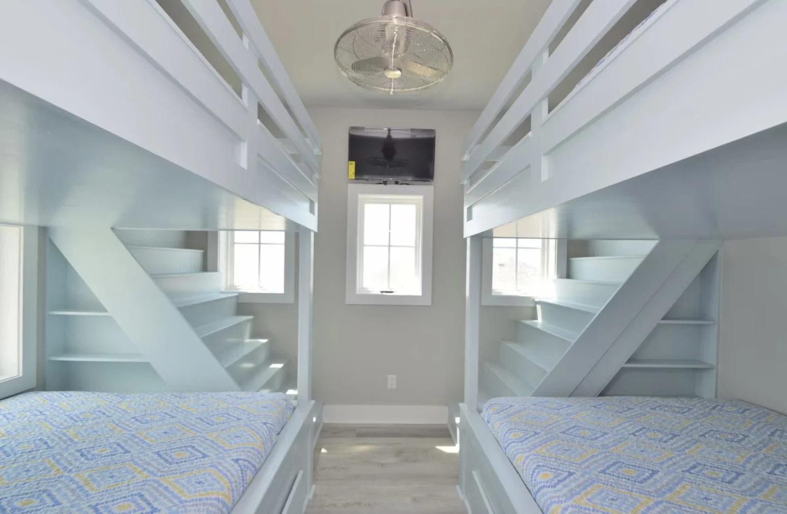 Upward view of a room with two sets of white bunk beds facing each other, each with a window in the background, a ceiling fan, and a mounted TV between the beds.