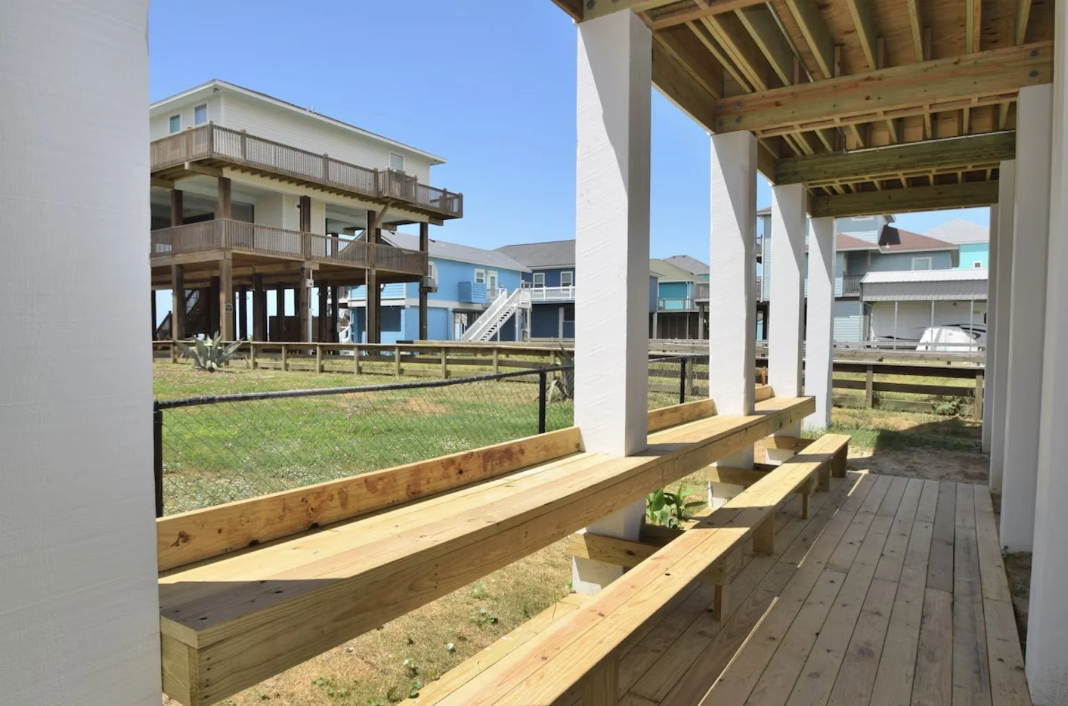 View from a covered porch with wooden seating, overlooking a backyard with a chain-link fence and neighboring beach-style houses with balconies.