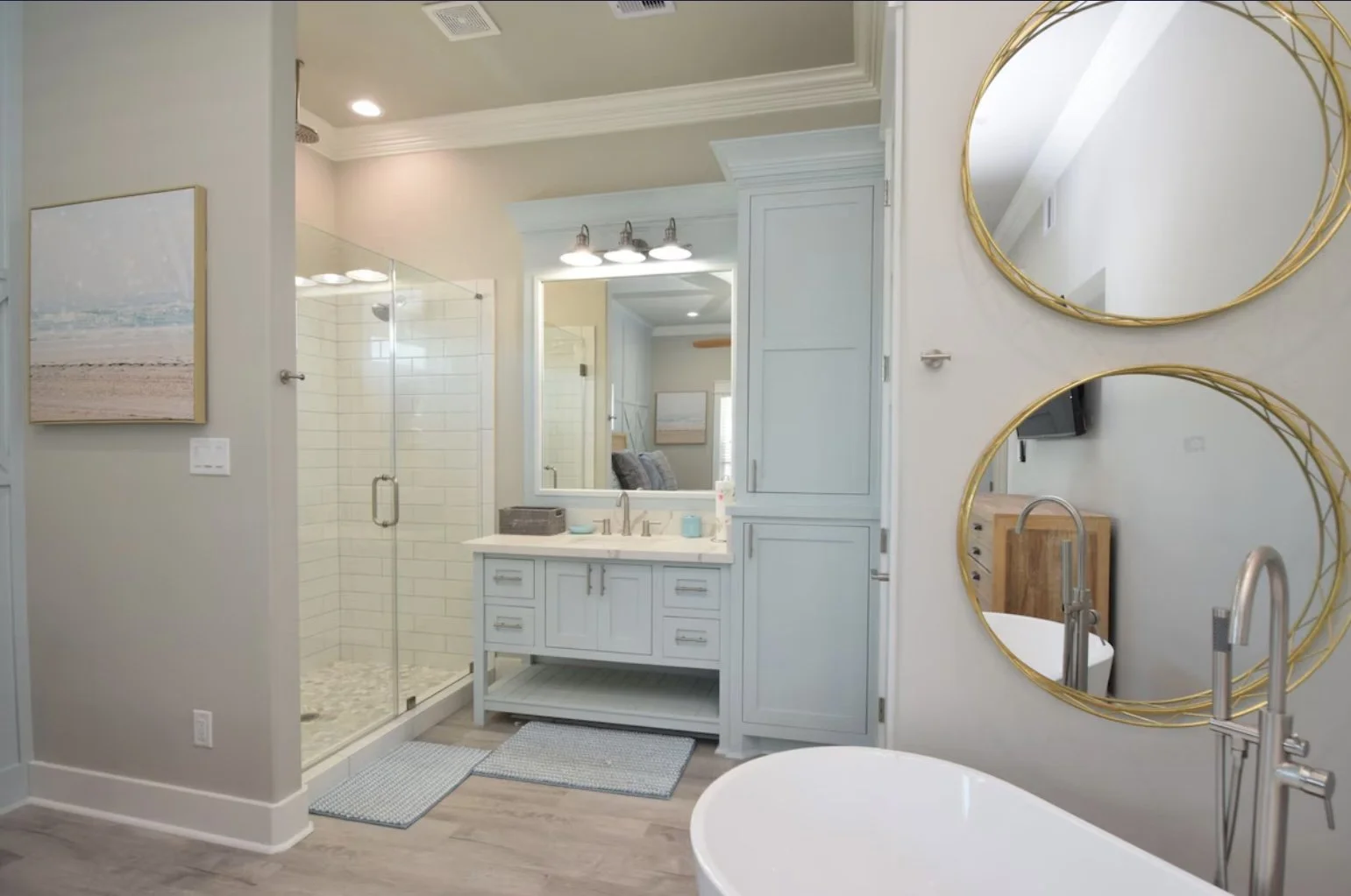 Bathroom with a glass shower, light blue cabinets, a large mirror, and two round gold-framed mirrors on the wall.