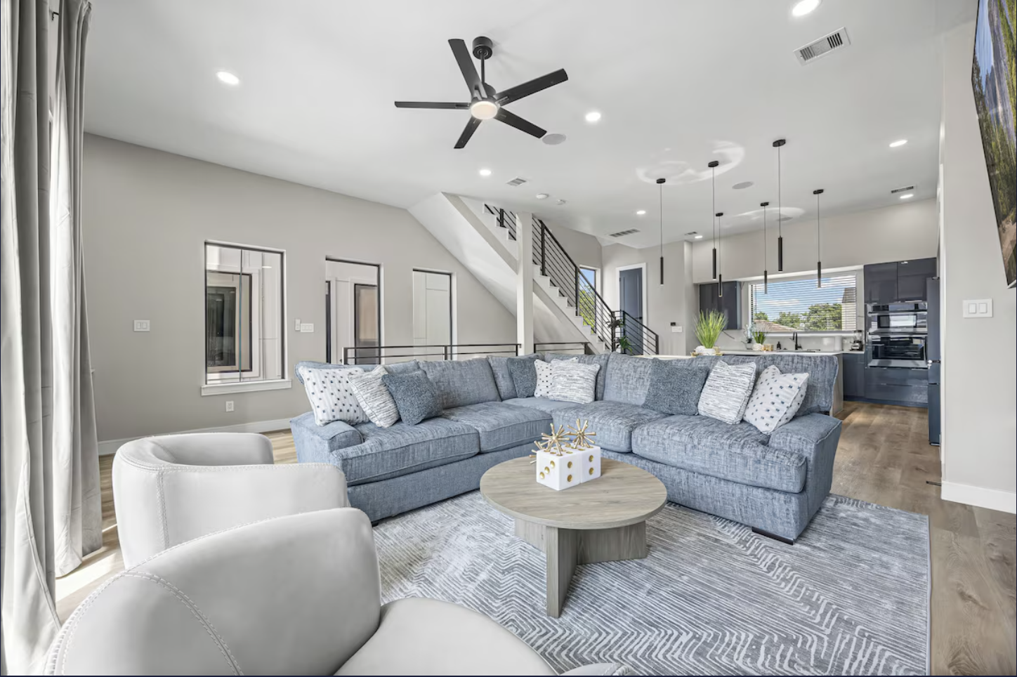 A modern open-concept living room with a large gray sectional sofa, white armchair, round wooden coffee table, ceiling fan, pendant lights, and a view of the kitchen with dark cabinetry and stainless steel appliances.