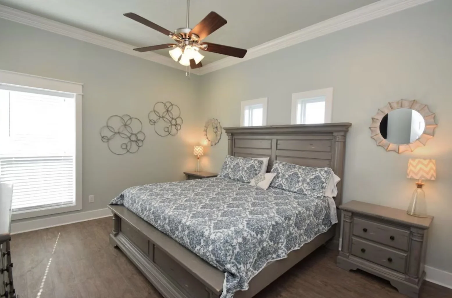 A bedroom with a large bed with a decorative headboard, patterned bedding, and pillows. There are two nightstands with lamps, small windows above the bed, a ceiling fan with lights, and wall decor on light-colored walls.