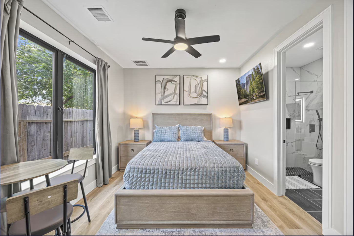 Bedroom with a bed, two nightstands with lamps, wall art, window with curtains, a ceiling fan, and a view of a bathroom with marble walls and shower.