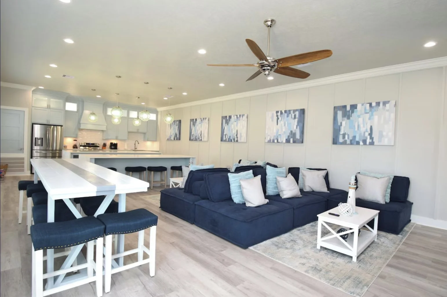 Open-concept living room with a navy blue sectional sofa, white coffee table, abstract artwork on the walls, and a kitchen with a large island, pendant lights, and stainless steel appliances.