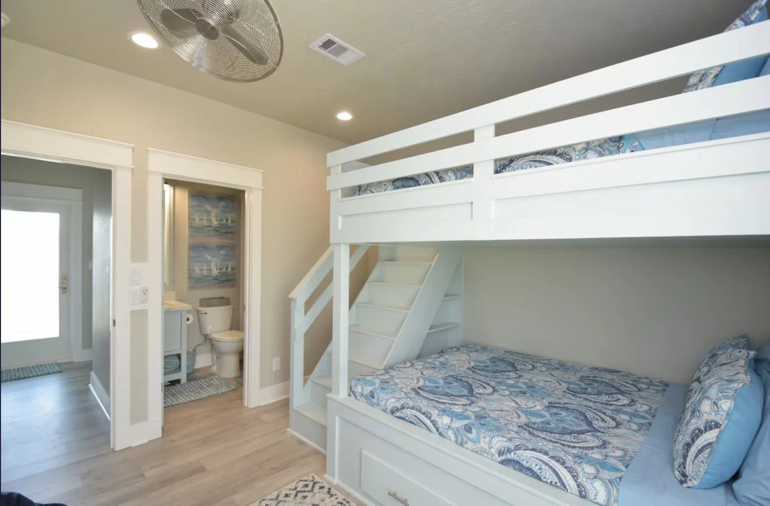 Interior of a cozy room with a bunk bed with blue and white bedding, a staircase, and a small bathroom with a toilet and ocean-themed wallpaper.