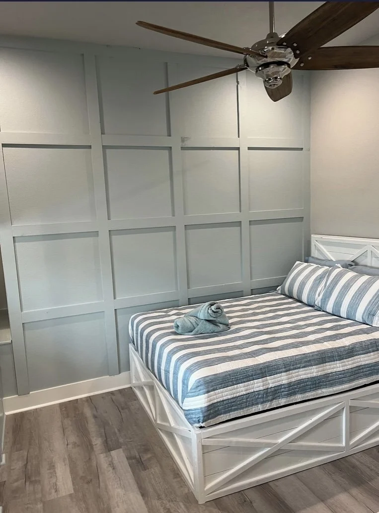 A bedroom with a white bed, striped bedding, and a wall with rectangular paneling. A ceiling fan with wooden blades is hanging from the ceiling.