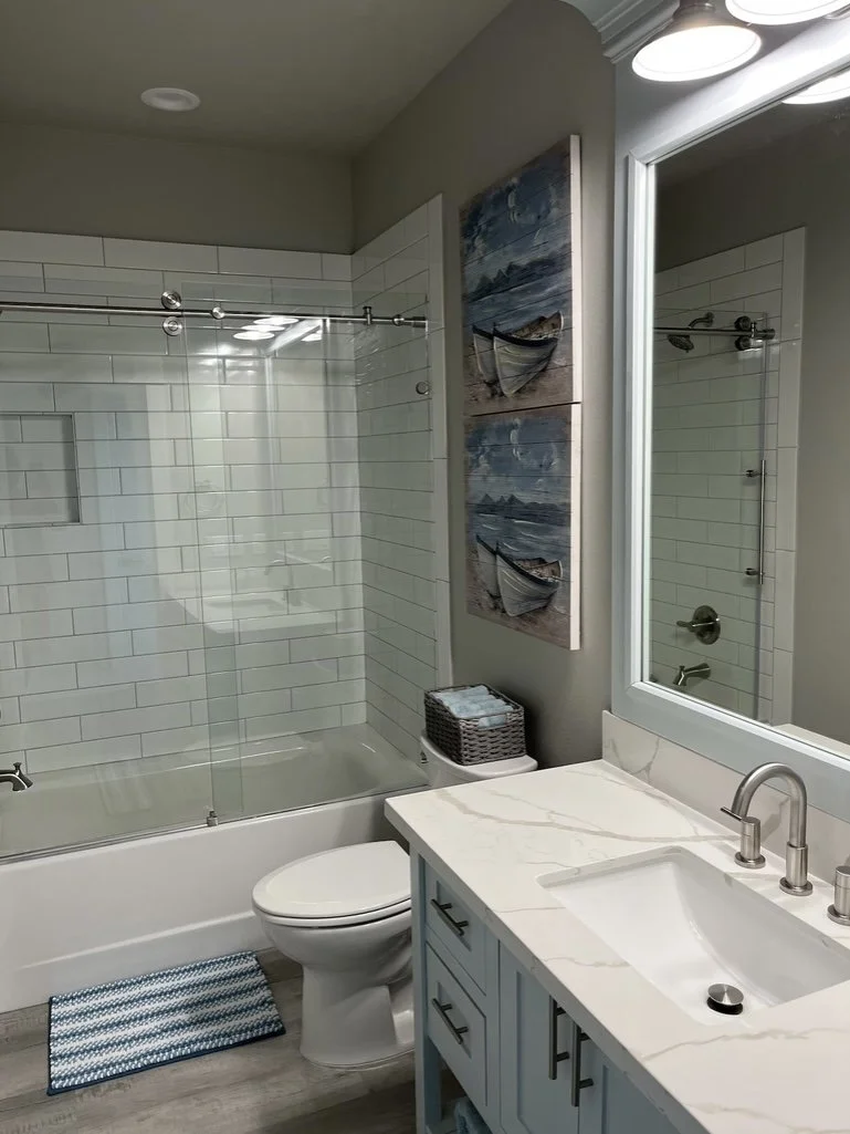 Bathroom with white tiled shower, toilet, vanity with marble countertop, framed mirror, wall art of boats, small basket, striped bath mat, and gray walls.