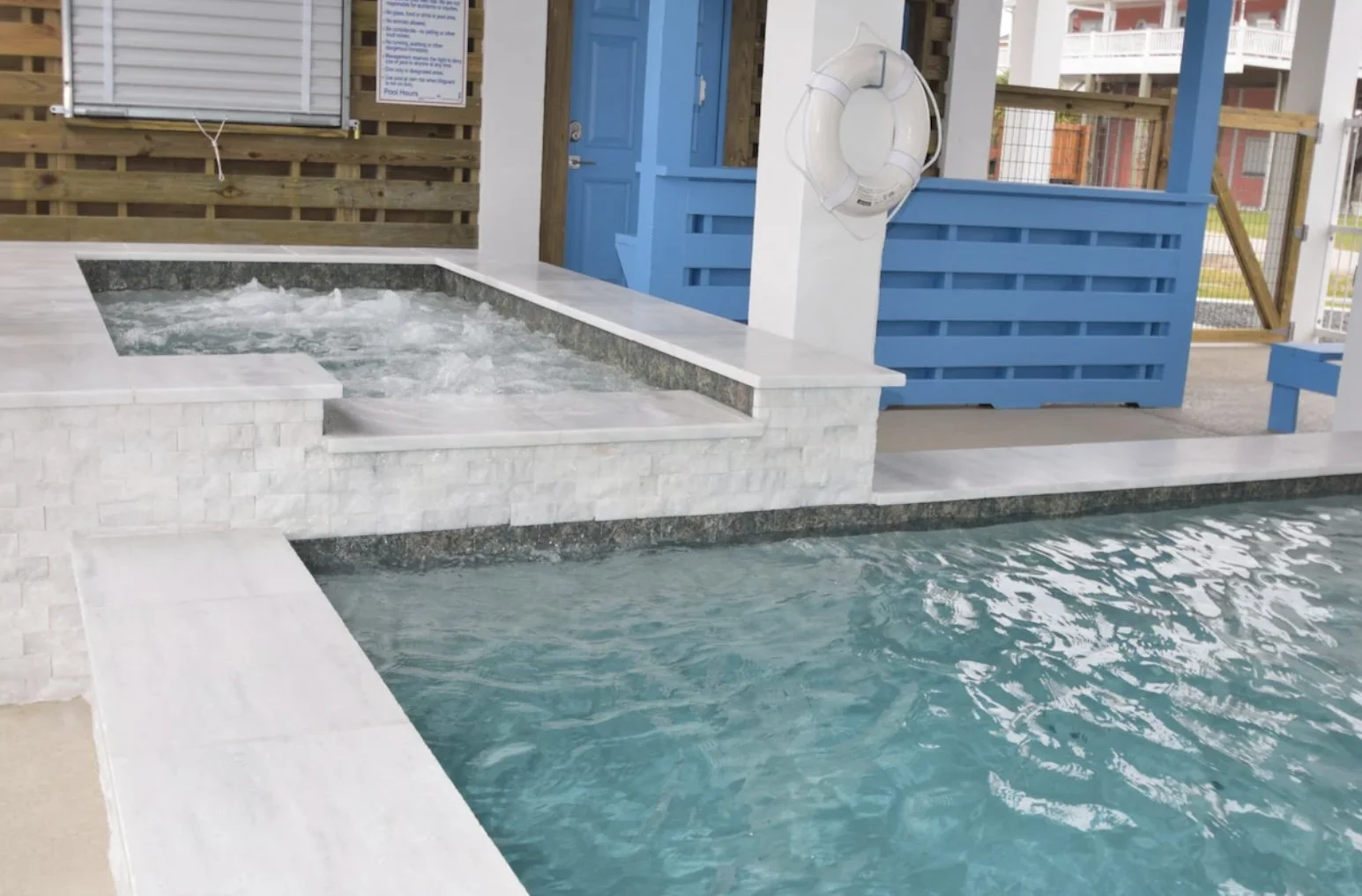 A small swimming pool with a bubbling hot tub adjacent to it, both surrounded by white tiles and a wooden fence, with a blue door and life preserver on the wall nearby.