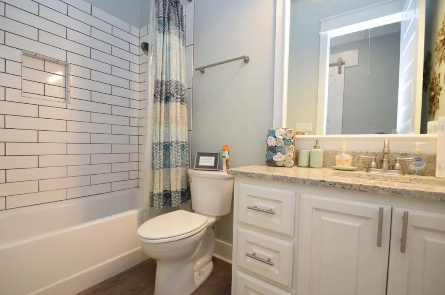 Bathroom with a tub and shower area, a toilet, and a vanity with a large mirror. The shower has white tile with black grout and a window in the tile wall. The vanity has a granite countertop, soap dispensers, and rolled towels. There is a framed sign