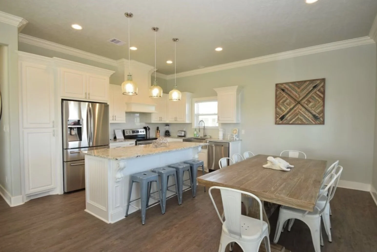 Kitchen with white cabinets, stainless steel refrigerator, and a wooden dining table with six white chairs. Pendant lights hang above the kitchen countertop with four metal stools. Wall art on light green wall.