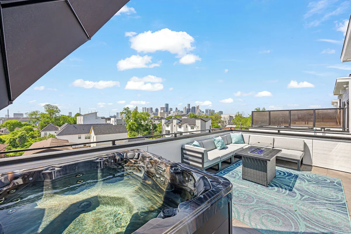 Outdoor rooftop patio with hot tub, sectional sofa, coffee table, area rug, umbrella, and city skyline in the distance under a blue sky with clouds.