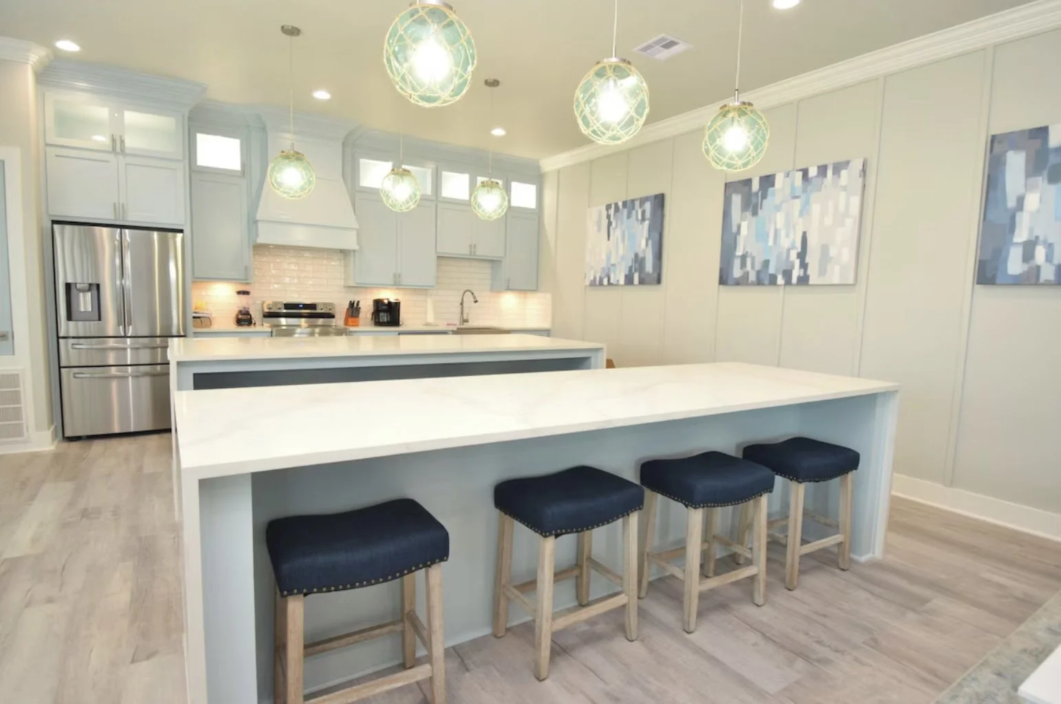 Modern kitchen with light blue cabinets, stainless steel refrigerator, white countertop island, and abstract blue and white artwork on the walls. Four black and wood bar stools are placed at the island, with pendant lights hanging above.