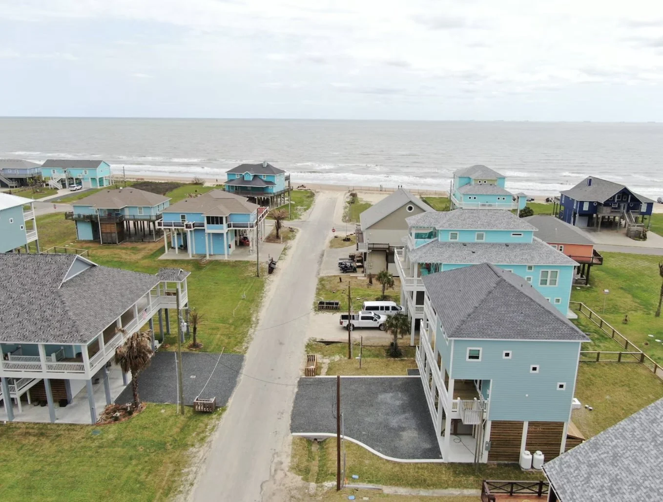 Aerial view of beach houses along a shoreline with a road in front, the ocean in the background, under a cloudy sky.