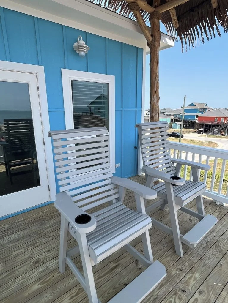Two white wooden rocking chairs on a wooden deck outside a blue beach house with a thatched roof overhang, with a view of beach houses in the background.