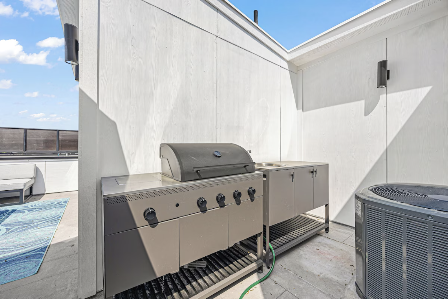Outdoor rooftop patio with a stainless steel gas grill, small sink, and air conditioning unit against white walls, under a blue sky with clouds.