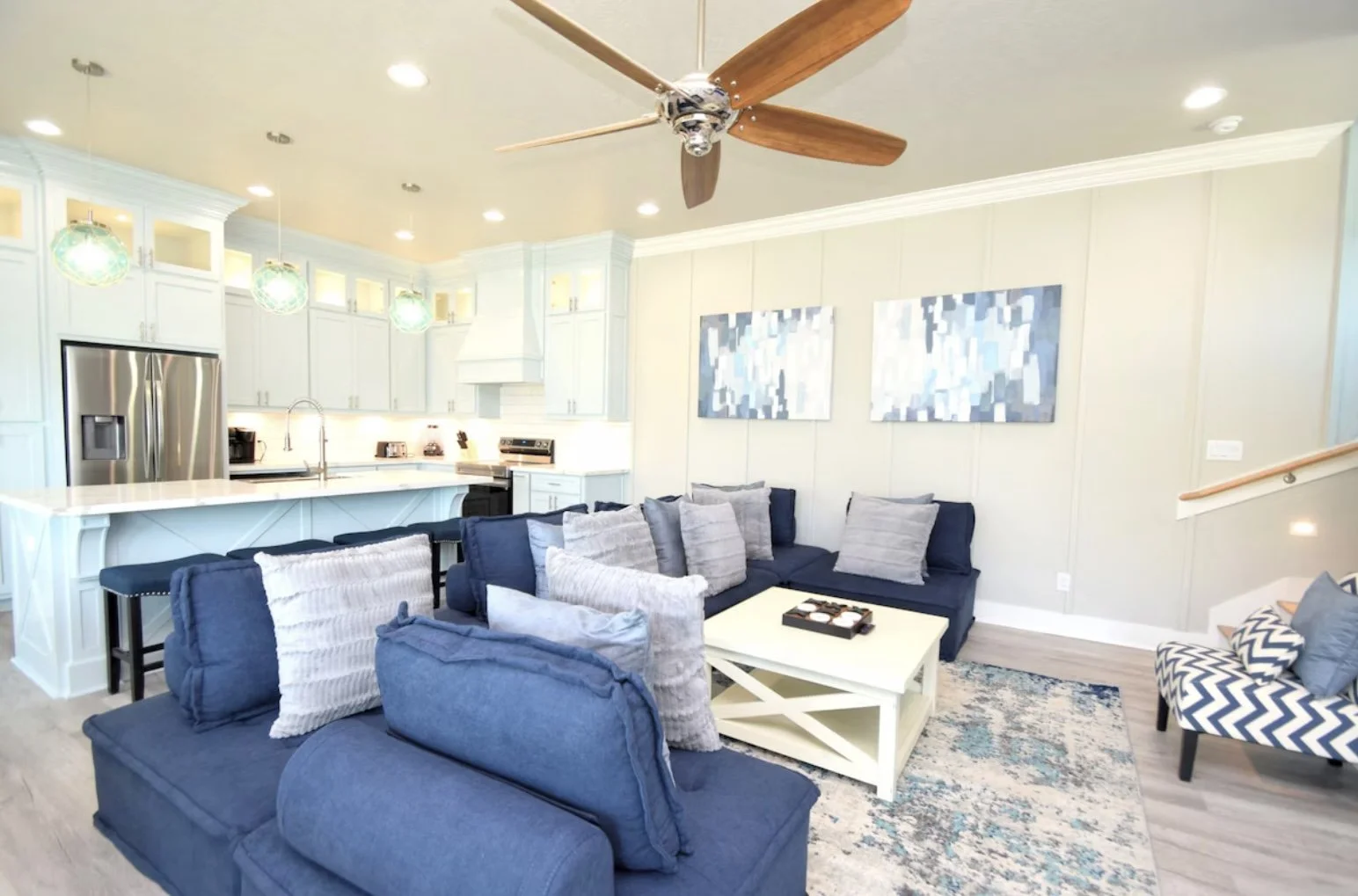 Living room with navy blue sofas, gray and patterned pillows, white coffee table, wall-mounted artwork, adjacent kitchen with white cabinets, stainless steel refrigerator, ceiling fan, and pendant lights.
