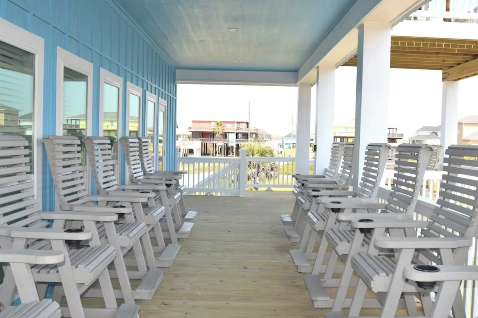 Beach-facing balcony with white wooden chairs arranged in two rows, light wood flooring, blue paneled wall, and neighboring houses in the background.