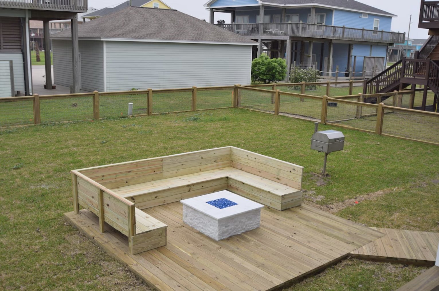 A backyard area with a newly built rectangular wooden deck that has an L-shaped built-in bench and a small fire pit table at the center. The yard is enclosed by a wooden fence, and there is a gray mailbox on a post nearby. In the background, there ar