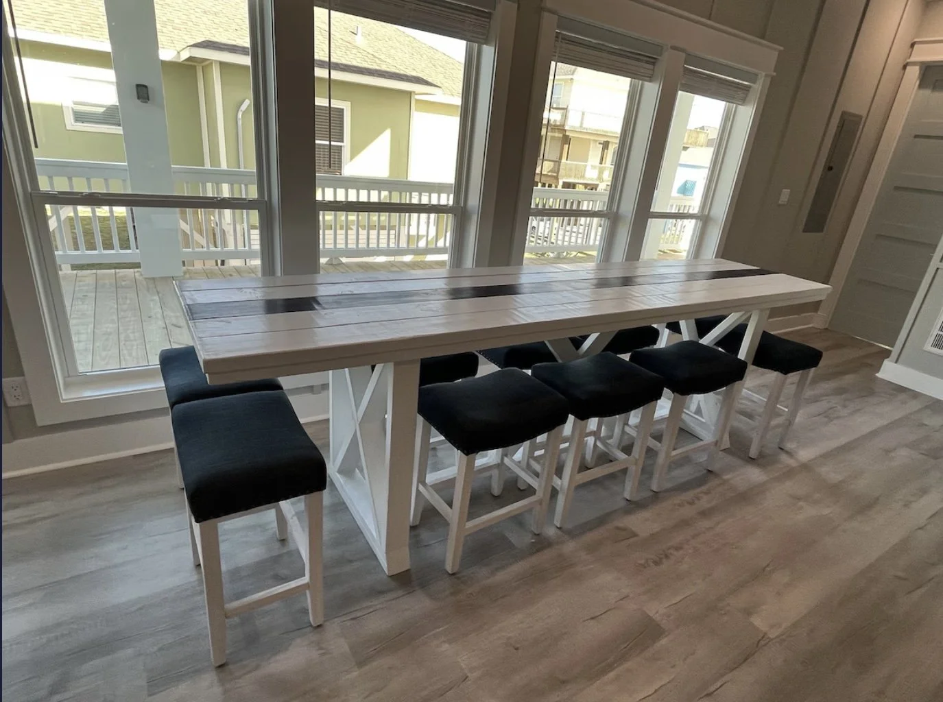 A wooden kitchen island with six black cushioned stools, situated in front of large windows overlooking a porch and neighboring houses.