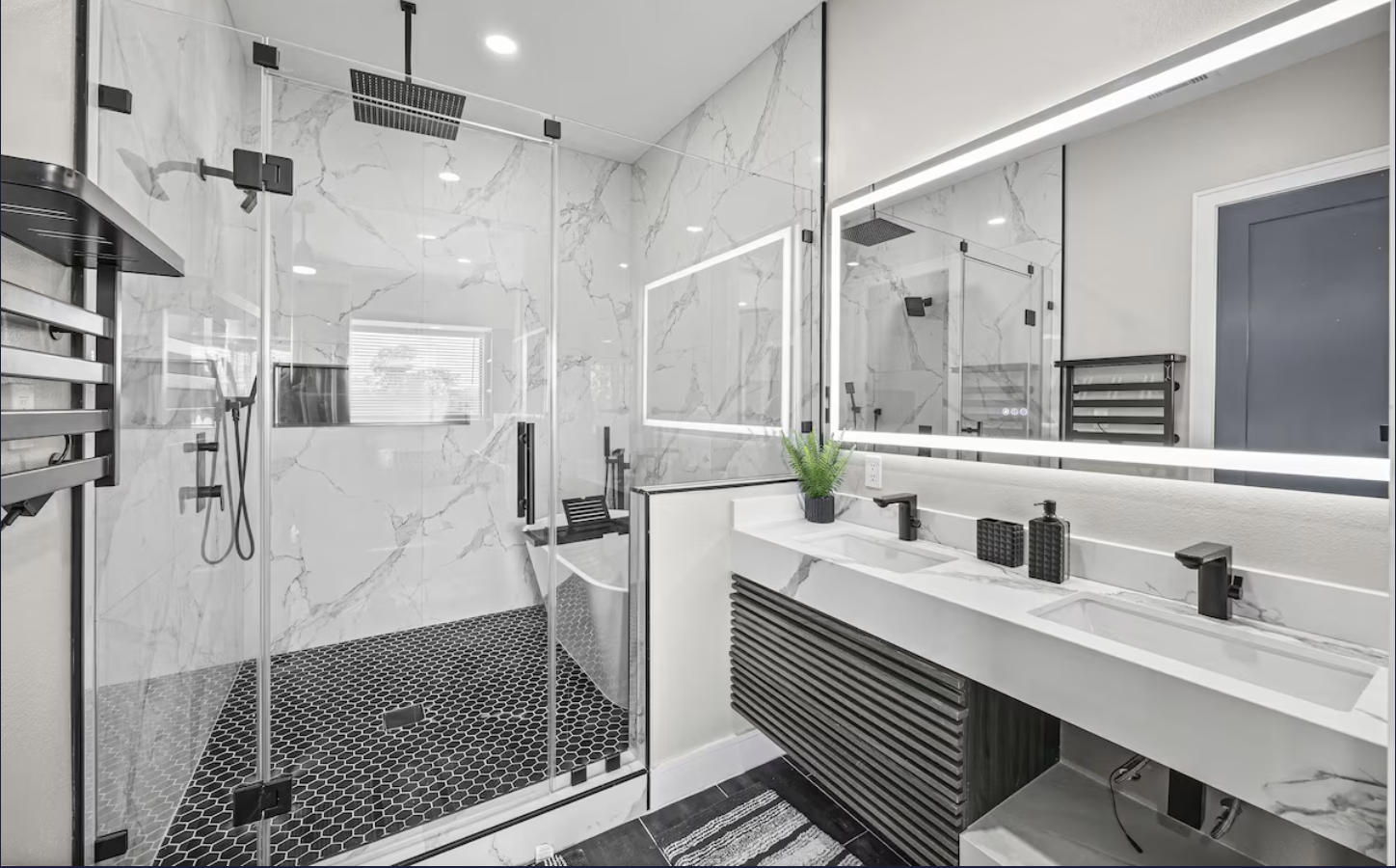 Modern bathroom with a walk-in glass shower featuring black fixtures, white marble walls, and a black hexagonal tile floor; a white double vanity with black fixtures, a large mirror with LED lighting, and a small potted plant.