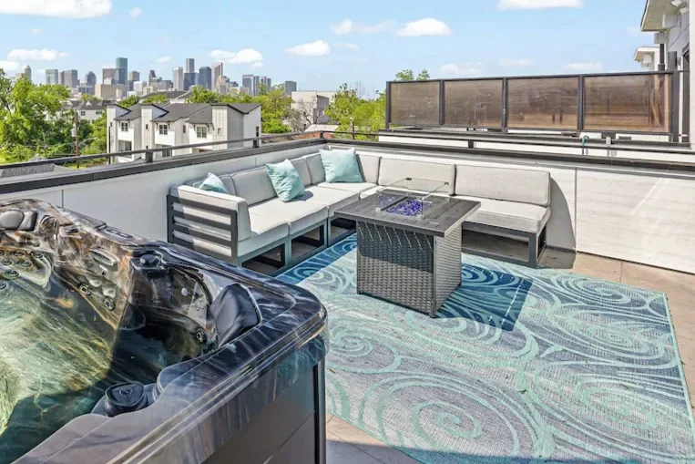 Outdoor rooftop deck with a sectional sofa, a coffee table, a hot tub, and a colorful outdoor rug, overlooking a city skyline with trees and residential buildings.