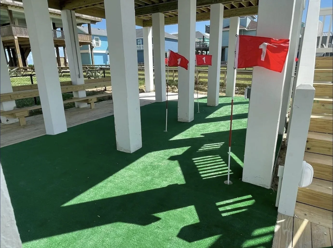Underneath a building structure with white support columns and green artificial grass, multiple red flags with white arrows and the number '1' are planted in the ground, creating a mini golf course or obstacle area.