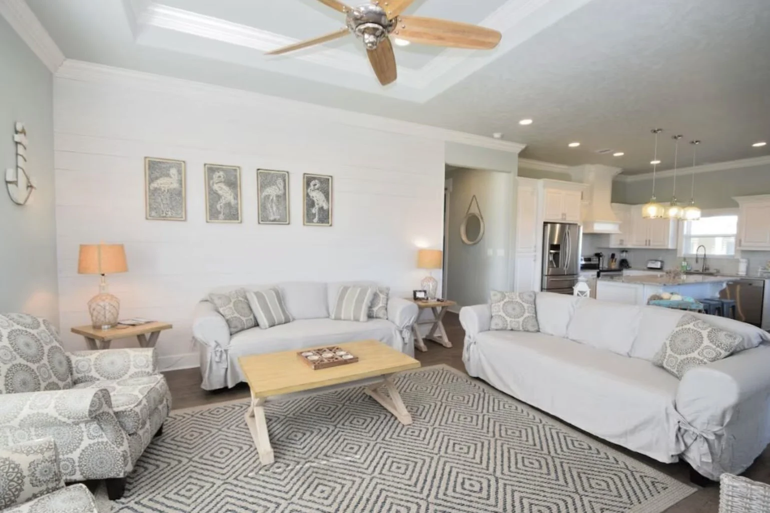 A bright living room with white sofas, a patterned armchair, a coffee table, and a rug. There are two table lamps, wall art of flamingos, and an open kitchen in the background with white cabinets and stainless steel appliances, including a refrigerat