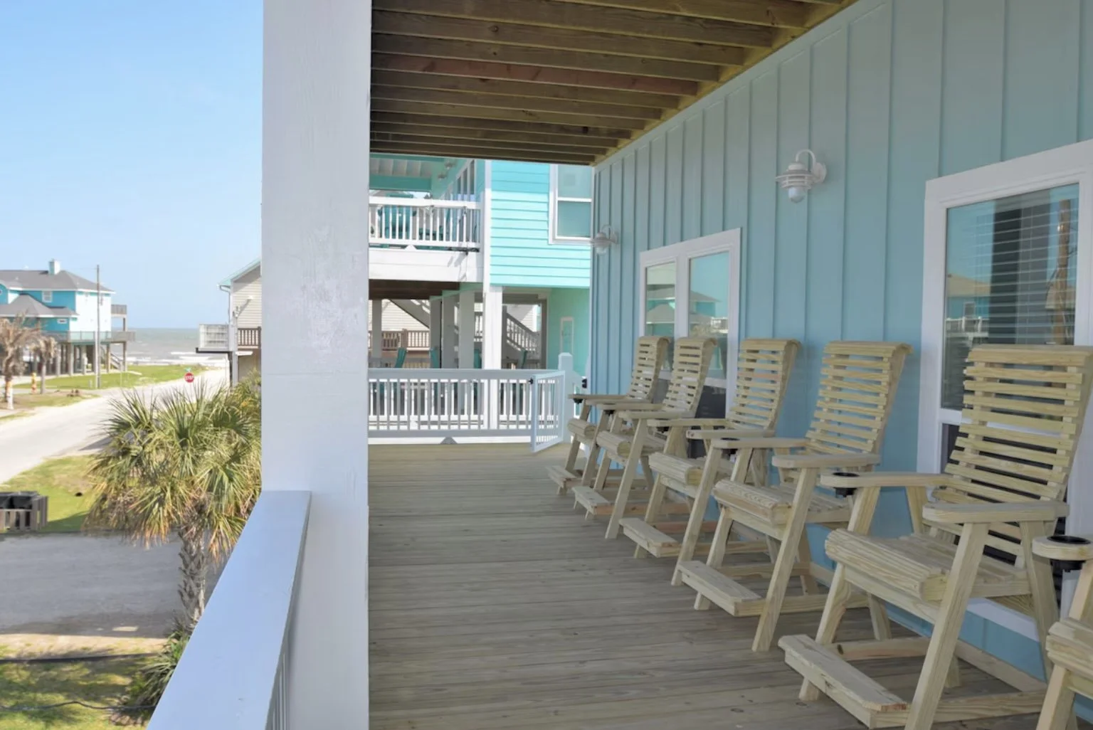 Beach house balcony with six wooden rocking chairs and ocean view in the background.