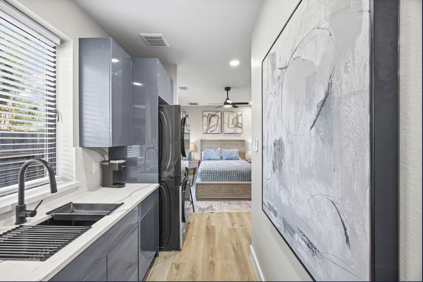 Modern laundry room with slate blue cabinets, white marble countertop, stainless steel sink, and front-loading washer and dryer. A large window with blinds allows natural light. Beyond the laundry area is a bedroom with a bed, artwork, and a ceiling 