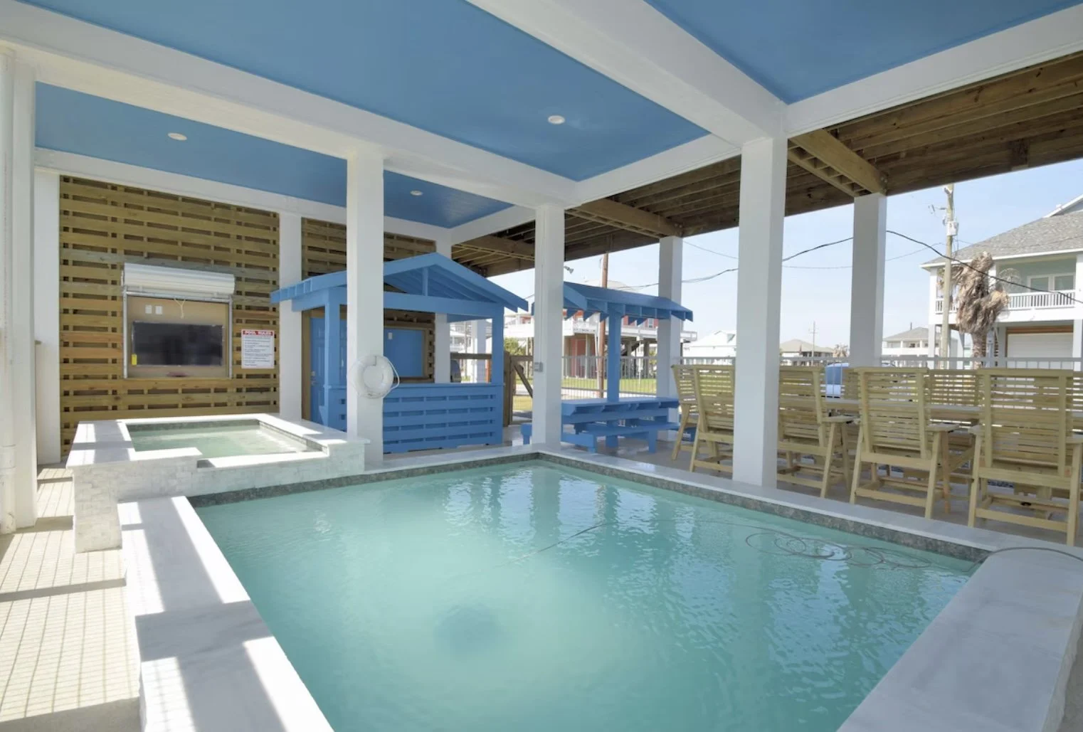 An outdoor pool area with a small hot tub, wooden seating, and a shaded playhouse, surrounded by a fence and neighboring houses.